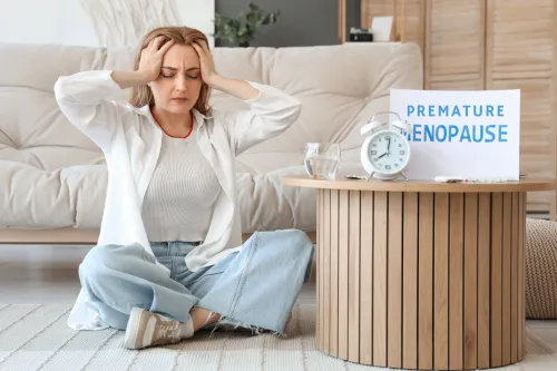 Middle-aged woman holding her head in discomfort while sitting next to a table labeled 'Premature Menopause,' symbolizing the emotional and physical symptoms of early menopause.