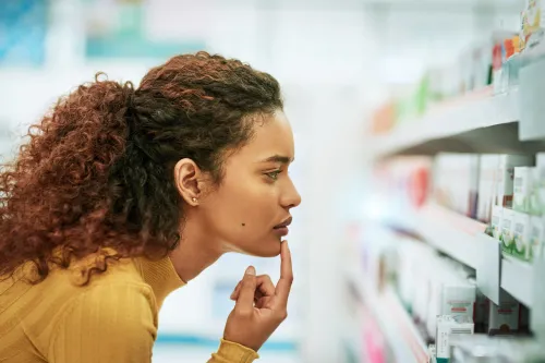 A woman in a mustard sweater thoughtfully examines medications on a pharmacy shelf, reflecting on whether to start thyroid medication.